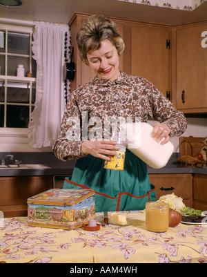 1960s SMILING WOMAN HOUSEWIFE MOTHER WEARING APRON IN KITCHEN POURING MILK INTO THERMOS FOR SCHOOL LUNCH Stock Photo