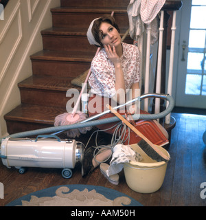 1960s 1970s TIRED BORED YOUNG WOMAN DAYDREAMING SITTING ON STAIRS SURROUNDED BY HOUSE CLEANING TOOLS Stock Photo
