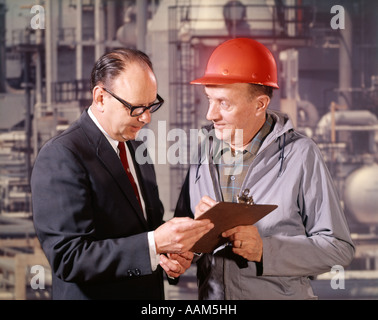 1960s EXECUTIVE AND ENGINEER WITH CLIPBOARD TALKING FACTORY REFINERY IN BACKGROUND OUTDOOR HARD HAT Stock Photo
