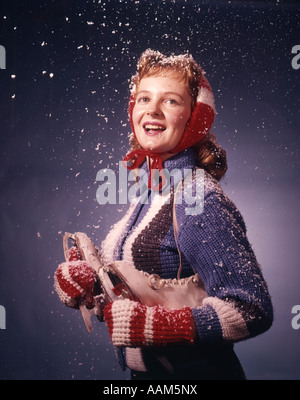 1960s SMILING YOUNG WOMAN WEARING RED MITTENS EAR MUFFS BLUE SWEATER HOLDING ICE SKATES IN FALLING SNOW Stock Photo