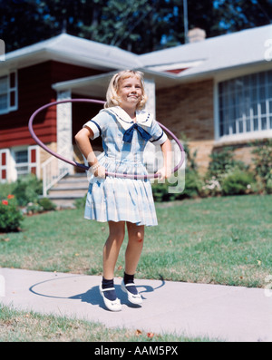 1950s 1960s LITTLE GIRL WITH HULA HOOP PLAYING OUTSIDE LAUGHING PURPLE ON SUBURBAN SIDEWALK IN SAILOR DRESS Stock Photo