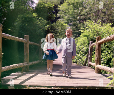 1960s LITTLE BOY AND GIRL HOLDING HANDS AND WALKING ACROSS WOODEN FOOTBRIDGE Stock Photo