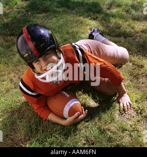 1970s FRECKLE FACED BOY FOOTBALL UNIFORM HELMET ORANGE TOP PINK PANTS LYING IN GRASS HOLDING FOOTBALL LOOK UP AT CAMERA Stock Photo