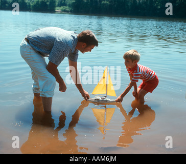 FATHER AND SON PUSHING A TOY SAILBOAT IN A LAKE RETRO 1970 1970s Stock Photo