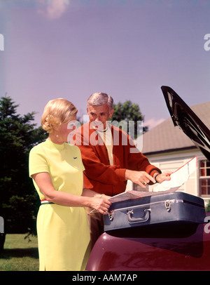 1960 1960s ELDERLY SENIOR COUPLE LOAD SUITCASES IN CAR TRUNK LOOKING AT MAP TRAVEL VACATION ROAD TRIP Stock Photo