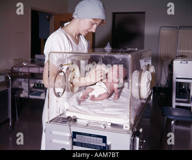 1960s WOMAN NURSE BOTTLE FEEDING INFANT BABY GIRL IN HOSPITAL NURSERY ISOLETTE PREMATURE INTENSIVE CARE Stock Photo