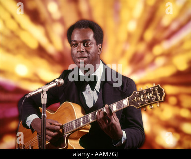 1970 1970s AFRICAN-AMERICAN MAN PLAYING GUITAR SINGING MICROPHONE TUXEDO GOLD LIGHTS BACKGROUND SINGER ENTERTAINER Stock Photo