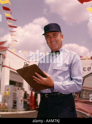 1960s MAN GAS SERVICE STATION MECHANIC HOLDING CHECK LIST ON CLIPBOARD BLUE UNIFORM HAT BLACK BOW TIE SMILING RETRO Stock Photo