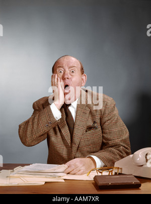 SHOCKED LOOKING BALDING EXECUTIVE WITH HAND TO CHEEK SITTING AT DESK WITH PAPERS MAN OFFICE UPSET 1960s Stock Photo