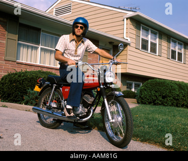 1970 1970s YOUNG MAN BLUE HELMET SITTING ON MOTORCYCLE IN DRIVEWAY SUBURBAN HOUSE BIKE MOTORCYCLES MEN Stock Photo
