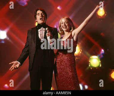 1970 1970s DUO MAN WOMAN COUPLE SINGING WOMAN RED DRESS GOWN MAN TUXEDO HOLDING MICROPHONE STAGE LIGHTS BACKGROUND Stock Photo