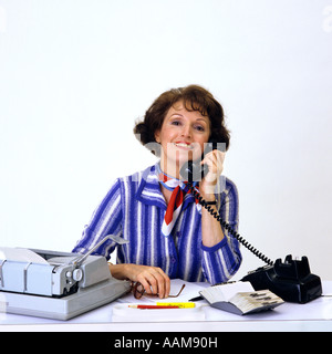 1980s BRUNETTE WOMAN ON BLACK TELEPHONE BLUE WHITE STRIPE BLOUSE SMILING AT DESK MANUAL TYPEWRITER ROLODEX SECRETARY Stock Photo