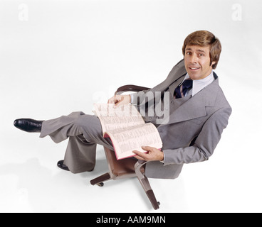 1970 1970s BUSINESSMAN GRAY 3 PIECE SUIT LOOKING UP BUSINESS BOOK DIRECTORY LAP SITTING IN OFFICE CHAIR MAN MEN RETRO Stock Photo
