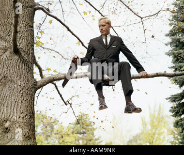 1960s MAN IN TREE SAWING OFF BRANCH HE IS SITTING ON ADAGE SYMBOLIC FOOLISH DANGER ONE Stock Photo