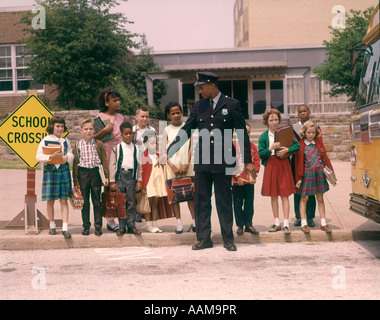 1960s AFRICAN AMERICAN MAN POLICEMAN PROTECTING BLACK AND WHITE SCHOOL CHILDREN CROSSING STREET IN FRONT OF SCHOOL BUS Stock Photo