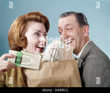 1960s COUPLE MAN WOMAN HOLDING SHOPPING BAG FULL OF MONEY CURRENCY SMILING HAPPY WEALTH DOLLARS WIN WINNINGS RETRO HUMOR Stock Photo
