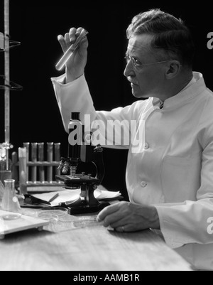 1920s 1930s 1940s SCIENTIST LAB TECHNICIAN IN WHITE COAT LOOKING AT TEST-TUBE IN FRONT OF MICROSCOPE Stock Photo