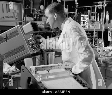 1960s MALE LABORATORY TECHNICIAN DRESSED IN A WHITE LAB COAT READING A OSCILLOSCOPE Stock Photo