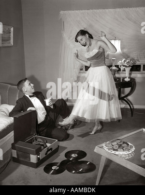 1950s 1960s TEEN COUPLE IN LIVING ROOM IN PROM DRESS & TUXEDO GUY SITTING ON FLOOR & GIRL DANCING TO MUSIC FROM A RECORD PLAYER Stock Photo