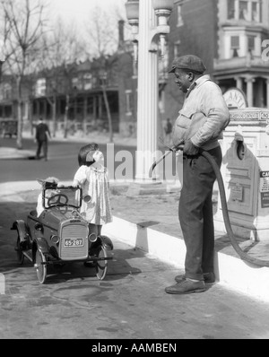 1920s SMILING LITTLE GIRL STANDING BESIDE HER PEDAL CAR ASKING FOR GASOLINE FROM AFRICAN AMERICAN GAS PUMP ATTENDANT Stock Photo