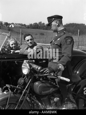 1930s COUPLE IN CONVERTIBLE COUPE STOPPED BY MOTORCYCLE COP CHECKING LICENSE OF DRIVER Stock Photo
