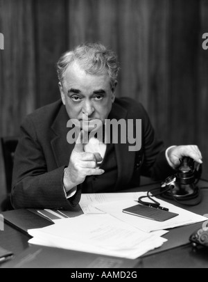1930s HEAD-ON SERIOUS SENIOR BUSINESSMAN BEHIND DESK HAND ON TELEPHONE POINTING Stock Photo