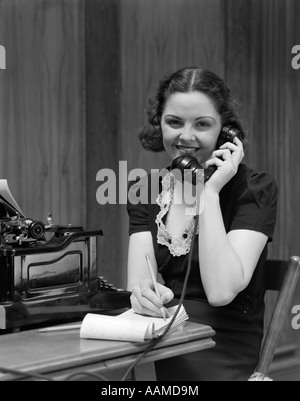 1930s SMILING YOUNG WOMAN RECEPTIONIST SECRETARY SITTING AT DESK IN OFFICE TALKING ON TELEPHONE Stock Photo