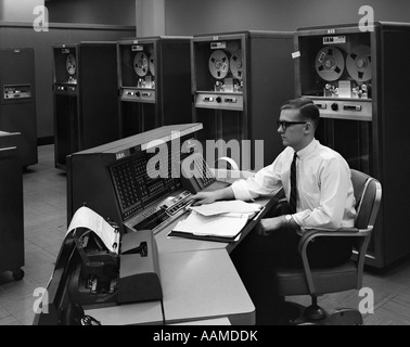 1960s SIDE VIEW OF MAN IN SHIRT TIE & THICK BLACK GLASSES WORKING WITH IBM DATA PROCESSING SYSTEM Stock Photo