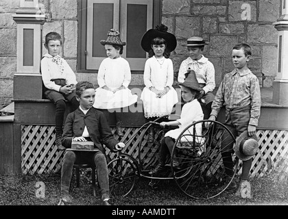 1890s 1900s TURN OF THE CENTURY GROUP OF SEVEN CHILDREN SITTING ON & AROUND PORCH ONE GIRL ON OLD FASHIONED TRICYCLE Stock Photo
