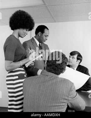 1970s GROUP OF TWO WHITE & ONE BLACK EXECUTIVES HAVING DISCUSSION WITH BLACK SECRETARY AT SIDE TAKING NOTES Stock Photo