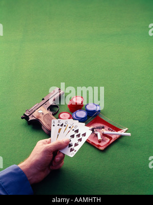 MAN HAND HOLDING PLAYING CARDS ON GREEN FELT CARD TABLE CIGARETTE POKER CHIPS ASH TRAY SEMI-AUTOMATIC PISTOL Stock Photo