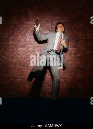 1970s 1980s MURDER MYSTERY MAN IN SUIT HOLDING GUN REVOLVER SHOT IN CHEST BRICK WALL PAIN AGONY CRIME DETECTIVE VIOLENCE Stock Photo