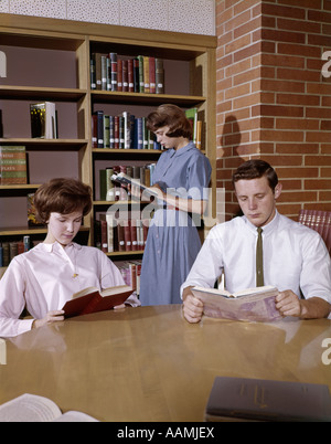 1960 1960s 3 STUDENTS BOY 2 GIRLS IN LIBRARY READING STUDY STUDYING HIGH SCHOOL EDUCATION RETRO SHELVES BOOKS Stock Photo