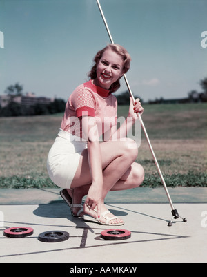 1950s SMILING BLONDE WOMAN KNEELING ON SHUFFLEBOARD HOLDING CUE POINT AT DISK WEAR WHITE SHORTS SHUFFLE GAME Stock Photo