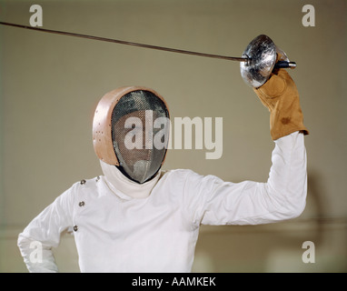 1960s MAN HOLDING FOIL SWORD EPEE ABOVE HIS HEAD WEARING HELMET PROTECTIVE GEAR WIRE FACE MASK ON GUARD TOUCHE FENCING Stock Photo