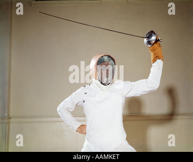 1960s MAN PROTECTIVE CLOTHING HELMET WIRE FACE MASK HOLDING WAVING FOIL EPEE SWORD ABOVE HEAD ON GUARD TOUCHE DUEL Stock Photo