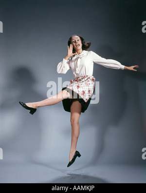 1960s YOUNG WOMAN WEARING APRON JUMPING INTO THE AIR AND SHOUTING HAPPY SYMBOLIC KICKING JOY TRIUMPH Stock Photo