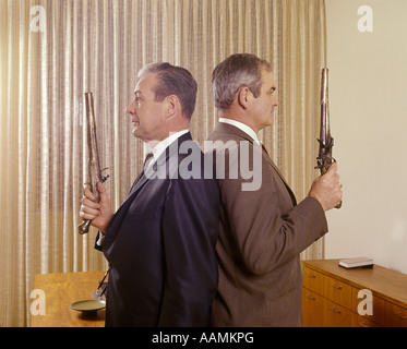 1960s 2 MIDDLE AGED BUSINESS MEN STAND BACK TO BACK HOLDING GUNS DUEL PISTOLS Stock Photo