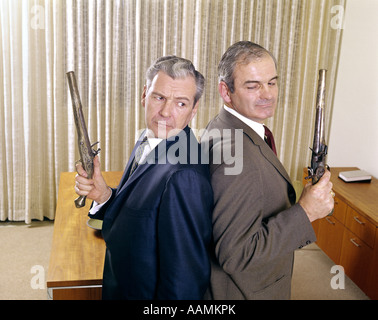 1960s 2 MIDDLE AGED BUSINESS MEN STAND BACK TO BACK HOLDING GUNS PISTOLS DUEL DUELLING ANGRY EXPRESSION ENEMY ENEMIES Stock Photo