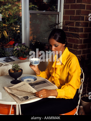 1970 1970s AFRICAN-AMERICAN WOMAN YELLOW BLOUSE SITTING TABLE BY WINDOW DRINKING MORNING COFFEE READING NEWSPAPER Stock Photo
