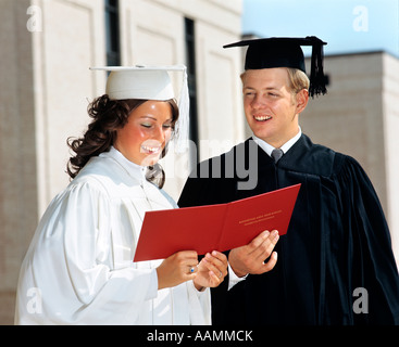 1970s TEEN COUPLE GIRL WHITE ROBE BOY IN BLACK ROBE MORTARBOARD HOLD RED DIPLOMA BETWEEN THEM HIGH SCHOOL SMILING PROUD Stock Photo