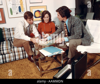 1970s RETRO INSURANCE SALESMAN SHOWING POLICY TO HUSBAND WIFE MAN WOMAN COUPLE SITTING ON COUCH IN LIVING ROOM Stock Photo