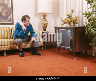 1960s MAN SITTING ON SOFA HOLDING ROLLED UP NEWSPAPER AND LOOKING AT TELEVISION LIVING ROOM INDOOR Stock Photo