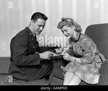 1940s A WARTIME ROMANCE COUPLE MAN IN MILITARY UNIFORM WOMAN IN PRINT DRESS SITTING IN CHAIR SMILING LOOKING AT PAPERS Stock Photo