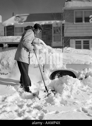 1950s MAN IN FRONT OF HOUSE SHOVELING SNOW FROM BURIED CAR Stock Photo