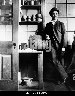1890s 1900s TURN OF THE CENTURY MAN IN SUIT & BOWLER LEANING ON WOODEN BARREL ON TABLE Stock Photo