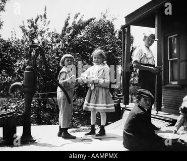 1890s 1900s TURN OF THE CENTURY GROUP OF CHILDREN STANDING ON FRONT PORCH TWO GIRLS PLAYING BY WATER PUMP Stock Photo