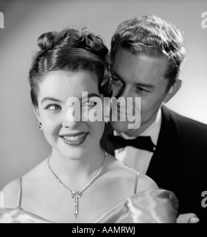 1950s 1960s SMILING HAPPY COUPLE WOMAN DIAMOND NECKLACE EARRINGS MAN BEHIND HER WEARING TUXEDO FORMAL EVENING ATTIRE Stock Photo