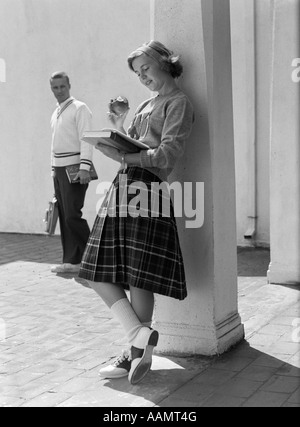 1950s 1960s TEEN SCHOOL GIRL PLAID SKIRT LEANING AGAINST COLUMN EATING APPLE READING BOOK AS BOY WALKS BY IN BACKGROUND Stock Photo