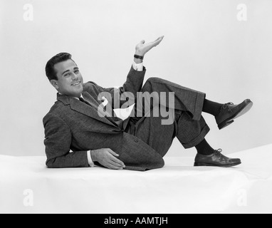 1950s MAN IN SUIT & TIE ON FLOOR LEANING BACK ON ELBOW WITH LEGS CROSSED & HAND IN AIR LOOKING AT CAMERA Stock Photo